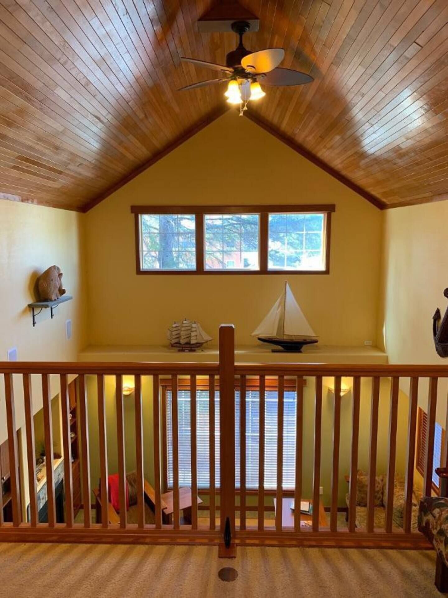 Une pièce chaleureuse avec un plafond en pente, des poutres en bois, un ventilateur de plafond et une fenêtre donnant sur les arbres.
