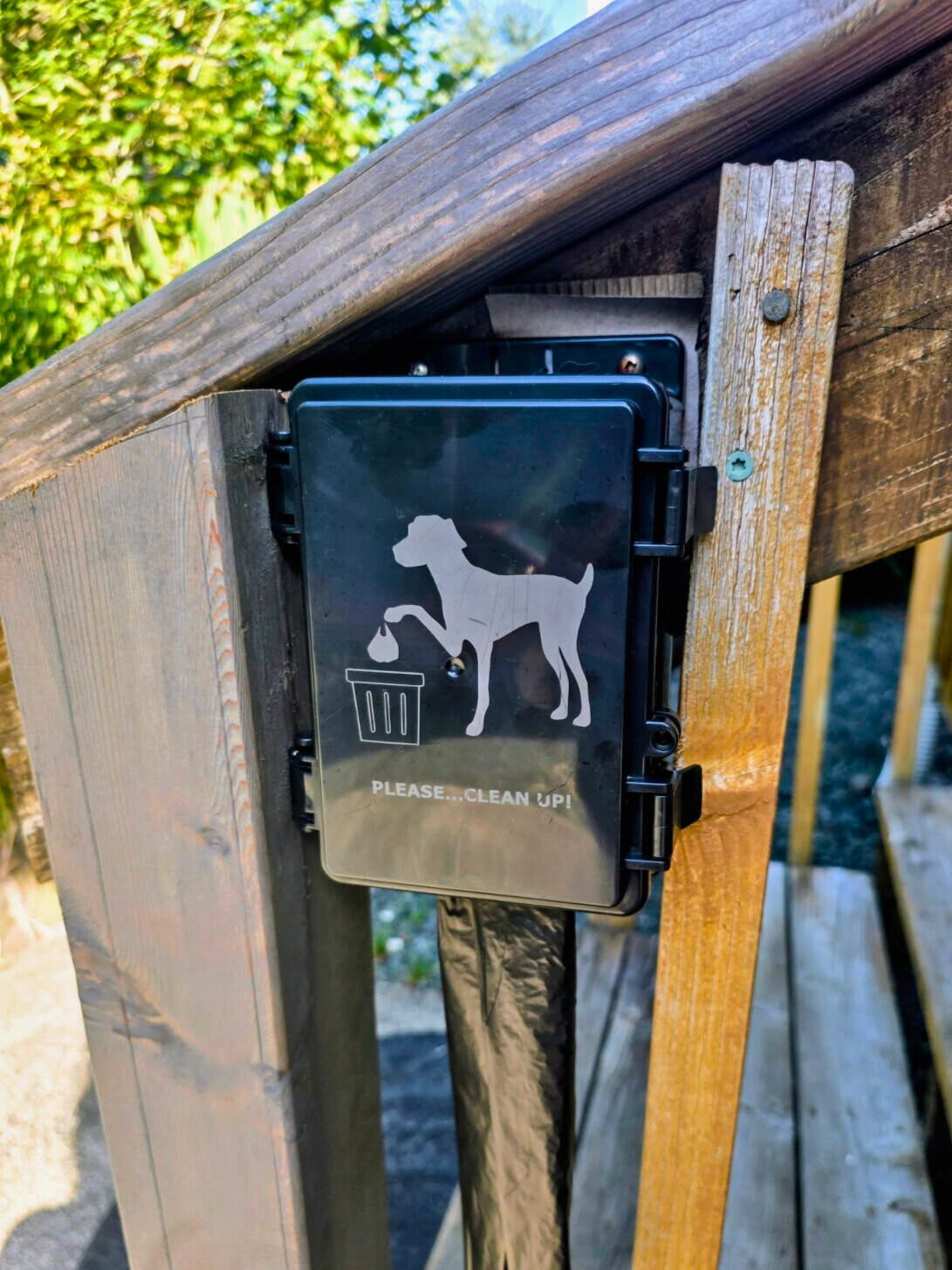 A metal trash bin attached to a wooden fence with a dog silhouette and a trash can icon, and a sign that says "PLEASE...CLEAN UP!"