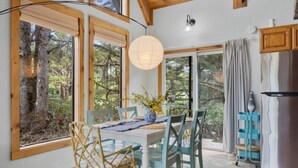A dining area with a table set for four, wooden chairs, a vase with flowers, and a view of trees through large windows.