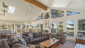 A spacious living room with large windows, a ceiling fan, and a wooden ceiling.