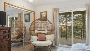 A bedroom with a large mirror, a wicker chair with cushions, a wooden dresser, and a view of a deck and trees.