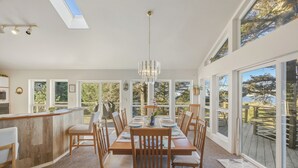 A dining area with a wooden table and chairs, a chandelier, and large windows offering a view of the outdoors.