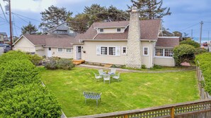 A two-story house with a stone chimney, a front porch, and a well-maintained lawn with outdoor furniture. - Susan’s Beach Cottage · Cozy Coastal Charm Just 1 Block from the Beach in the Heart of Manzanita (Manzanita)
