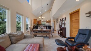 A spacious living room with a large sofa, a coffee table, and a dining area with a wooden table and chairs. There is a black leather chair and a bookshelf in the room.