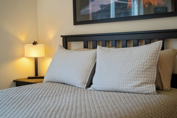 Una cama bien hecha con colcha acolchada, dos almohadas, una lámpara de noche y un cuadro en la pared.