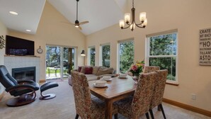 A spacious living room with a fireplace, a dining area with a wooden table and patterned chairs, and large windows offering a view of the outdoors.
