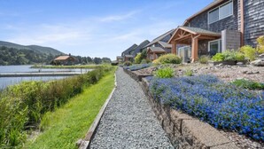 A row of houses with a gravel path and flower beds, a lake, and a hilly landscape in the background.