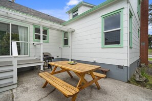 Une table de pique-nique en bois, avec des bancs, placée devant une maison blanche aux bordures vertes et dotée d’une cheminée en briques.