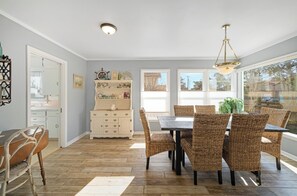 Un coin repas avec une table en bois et des chaises en osier, un meuble blanc agrémenté d’objets décoratifs, et une vue dégagée grâce à de grandes fenêtres.