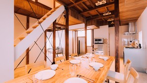 Un coin repas avec une table en bois dressée pour quatre personnes, un escalier et une cuisine en arrière-plan.