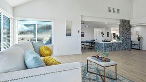 Un salon spacieux avec un canapé blanc, une table basse et une vue sur un coin cuisine agrémenté d’un mur en pierre.