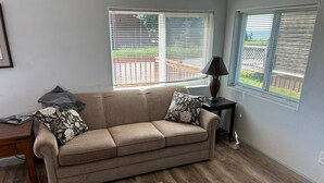 A living room with a beige sofa, two patterned pillows, a small wooden side table, a lamp, and a window with blinds.