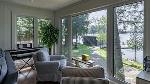 A living room with a piano, striped chairs, a large window with a lake view, and a small table with a radio. - 4bed, 4bath, South exp, prestigious area of Royal Muskoka Island w/road access (Minett)