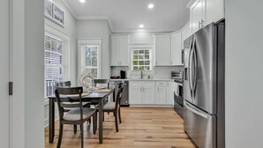 A modern kitchen with white cabinetry, a stainless steel refrigerator, and a dining area with a wooden table and chairs. - Ocean Lakes New Build, Steps to Beach and Balcony! (Myrtle Beach)