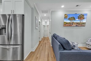 A modern kitchen with a stainless steel refrigerator, a wooden floor, and a grey sofa with patterned cushions. - Ocean Lakes New Build, Steps to Beach and Balcony! (Myrtle Beach)