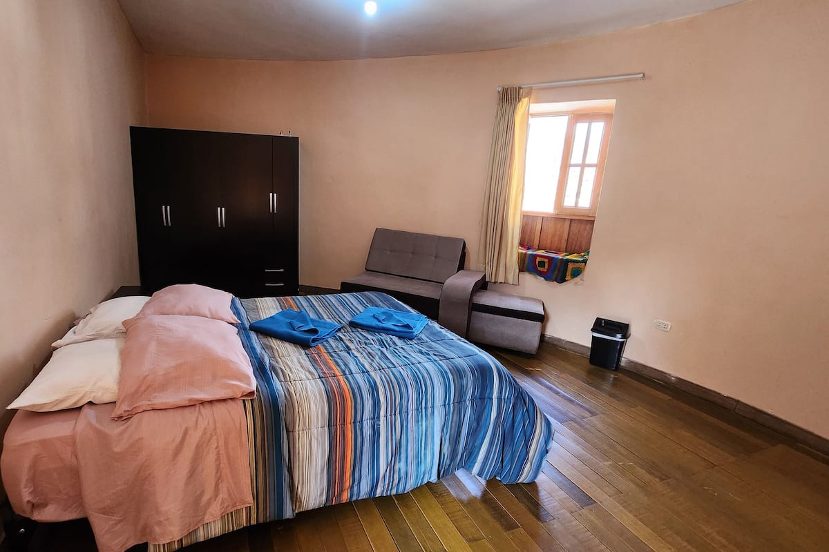 A bedroom with a bed, a wardrobe, a bench, a window with curtains, and a trash bin.