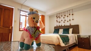 A plush llama toy on a glass table in a hotel room with a bed, wooden door, and a wall with hanging decorations. - Munay Cusco (Cusco)
