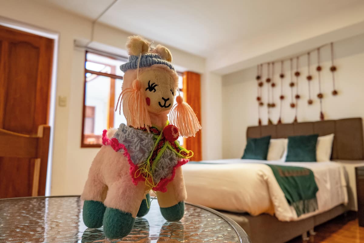 A plush llama toy on a glass table in a hotel room with a bed, wooden door, and a wall with hanging decorations.