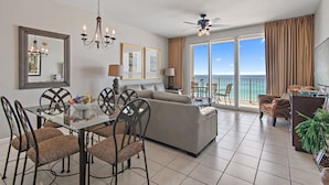 A spacious living area with a dining table, chairs, a sofa, and a balcony with a view of the ocean.