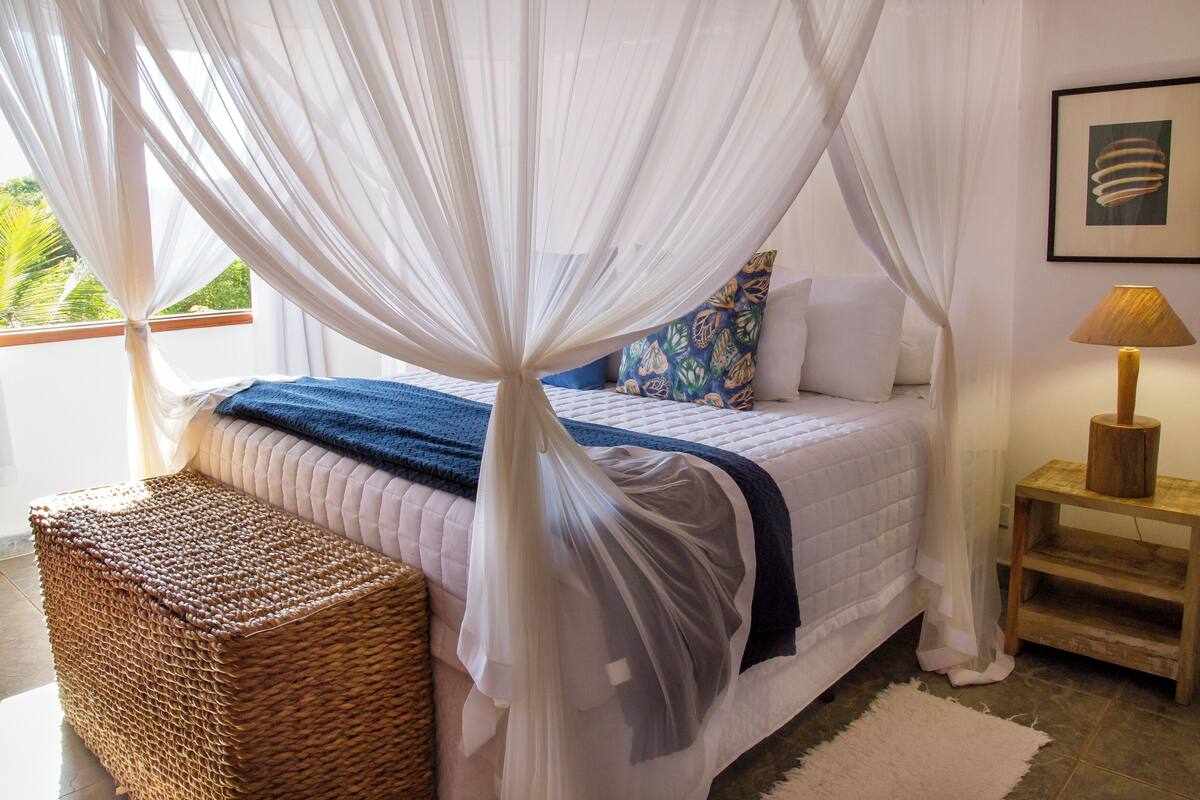 A bedroom with a bed under a canopy, a bedside table with a lamp, a wicker bench, and a view of greenery outside.