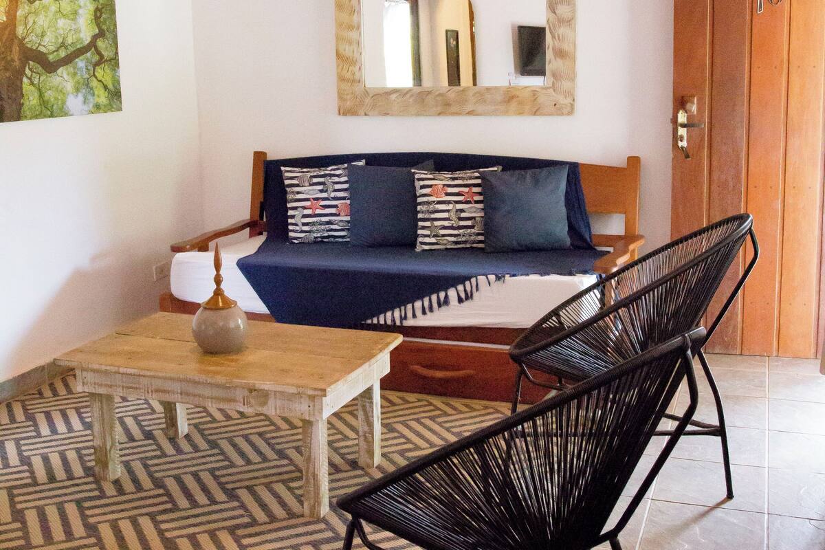 A room with a sofa, a chair, a table, a hanging light fixture, and a mirror on the wall.