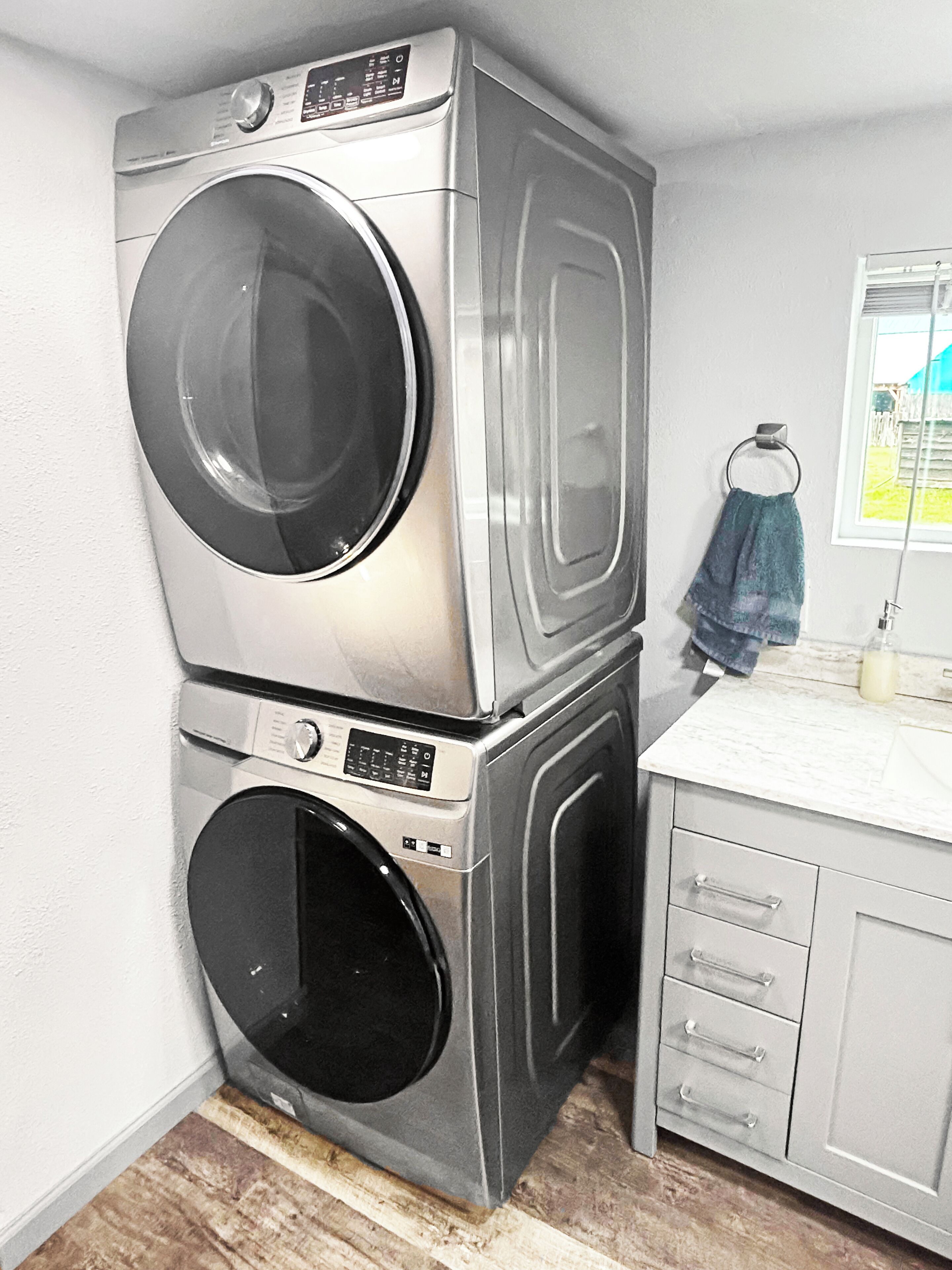 A compact laundry room with stacked washer and dryer units, a small countertop, and a window with a view of a green field.