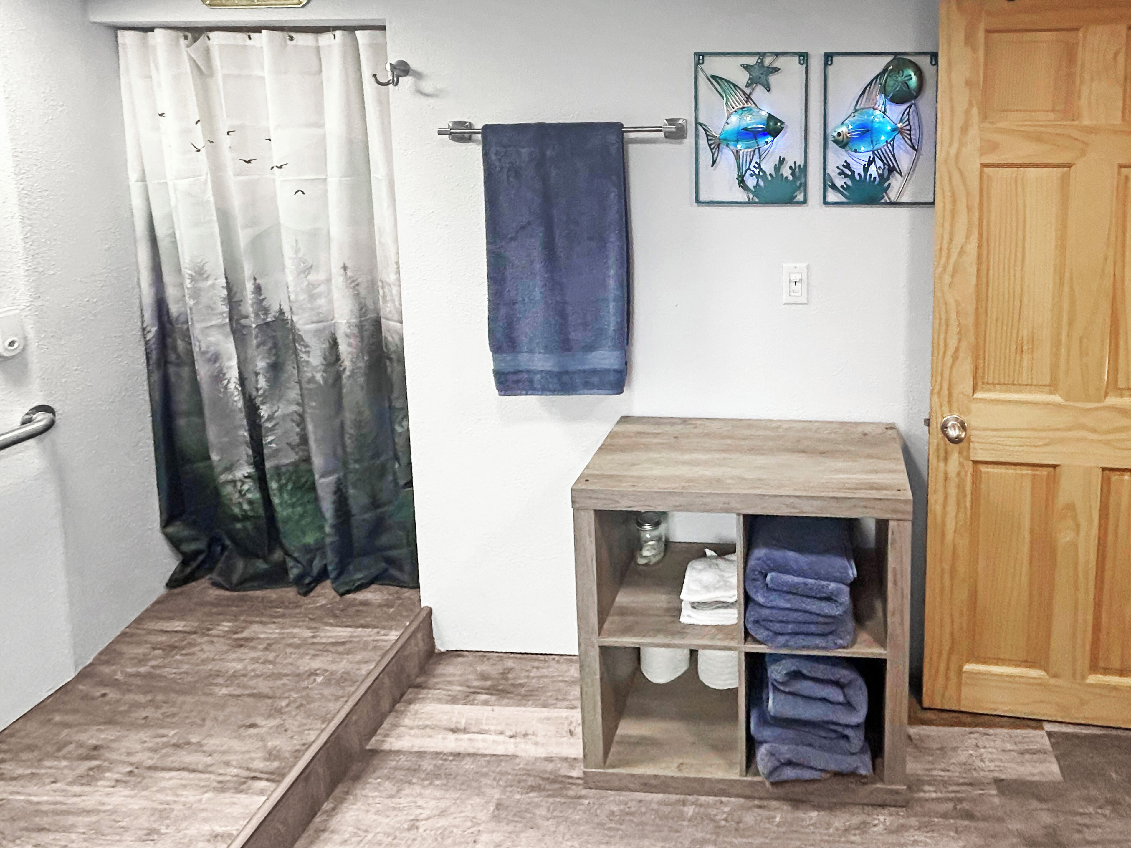 Un baño con cortina de ducha, un perchero, un estante de madera con toallas y dos adornos de peces colgados en la pared.