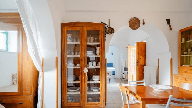 A dining area with a wooden table and chairs, a cabinet with glass doors, and a view into another room.
