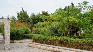 A courtyard with stone paving, a stone pillar, and various green shrubs and trees.