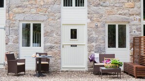 A stone building with a white door labeled "Jasper's Suite," a small table with chairs, and a wicker chair with a basket of flowers.