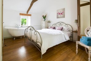 A bedroom with a bed, a vanity with a mirror, a window, a bathtub, and a wooden floor. - Juniper`s Stable. (Camborne)
