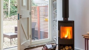 A room with a stone wall, a black wood-burning stove, a door leading outside, and a small table with a chair. - Tack Room a luxury Poldark retreat (Camborne)