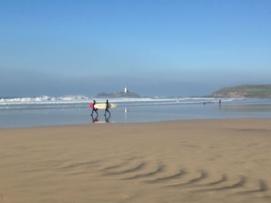 A beach with a lighthouse in the distance, sandy shore, and people walking with surfboards.