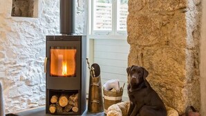 A cozy room with a stone wall, a wood-burning stove, and a dog resting in a bed. - The Hayloft - A Romantic Boutique Retreat (Camborne)