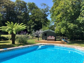 Ein Schwimmbad mit Holzterrasse, umgeben von üppiger Vegetation und einem kleinen Nebengebäude.