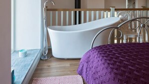A bedroom with a large window, a white bathtub, a wooden ceiling, and a metal bed frame. - The Hayloft - A Romantic Boutique Retreat (Camborne)