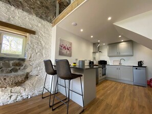 A kitchen with a stone wall, wooden flooring, and a bar area with stools. - Bramble’s Cottage (Cornwall)