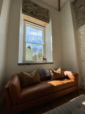 A room with a brown leather sofa, a stone wall, and a window with a view of a tree and a deer. - Bramble’s Cottage (Cornwall)