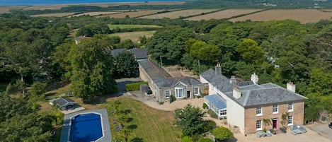 A large house with a swimming pool, surrounded by lush greenery and fields.