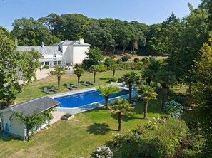 A large swimming pool surrounded by palm trees and a white building with a glass conservatory. - Bramble’s Cottage (Cornwall)