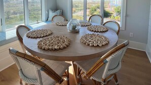 A dining area with a round table, wicker chairs, a decorative chandelier, and large windows offering a view of the outdoors. - 1 MINUTUE WALK TO BEACH FROM BHI BEACH FRONT HOME (Bald Head Island)
