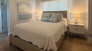A bedroom with a large bed, a ceiling fan, a window with blinds, a bedside table with a lamp, and a painting on the wall. - 1 MINUTUE WALK TO BEACH FROM BHI BEACH FRONT HOME (Bald Head Island)