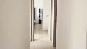 A narrow hallway with white walls and a tiled floor, leading to a room with a bed and a mirror.