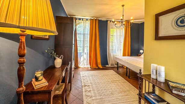 Ein gemütliches Zimmer mit Holztisch, Lampe, Bett und einem Fenster mit Vorhängen.