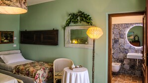 A room with a bed, a chair, a table, a lamp, a mirror, and a bathroom with patterned tiles.
