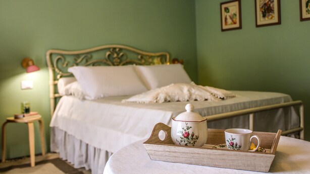 A neatly made bed with a white comforter and pillows, a bedside table with a lamp, and a tray with a teapot and cup on a round table.