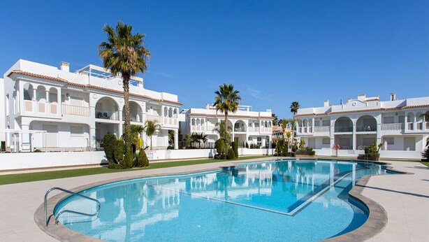 A swimming pool surrounded by white buildings with arched windows and balconies, palm trees, and a clear blue sky. - CH Allegra 135 Doña Pepa (Ciudad Quesada) (Ciudad Quesada)