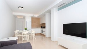 A modern living room with a grey sofa, a dining area with a glass table and chairs, a kitchen with white cabinets, and a large flat-screen TV mounted on the wall. - CH Allegra 135 Doña Pepa (Ciudad Quesada) (Ciudad Quesada)
