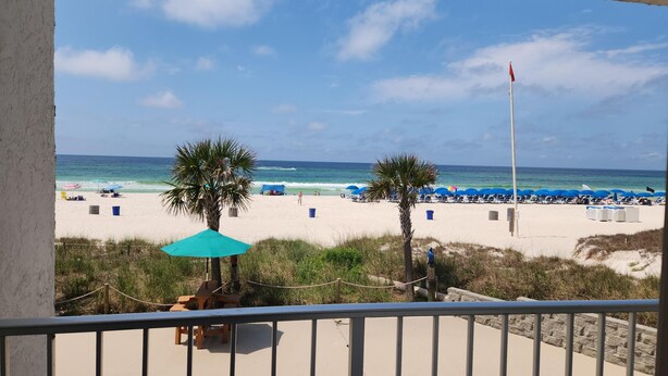 A beach view from a balcony with a railing, palm trees, and beach umbrellas. - Ambassador 128- Gulf Front - Pet Friendly (Panama City Beach)