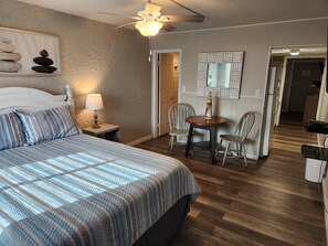 Une chambre à coucher avec un lit, une table de chevet avec une lampe, un ventilateur de plafond, une table à manger avec des chaises et un miroir au mur.